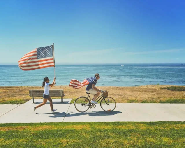 A person cycles along a seaside path, draped in an American flag. Another runs alongside, waving a large flag against a clear blue sky.