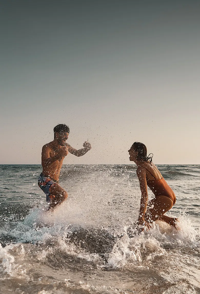 A man and woman joyfully play in ocean waves, splashing water under a clear sky during sunset.