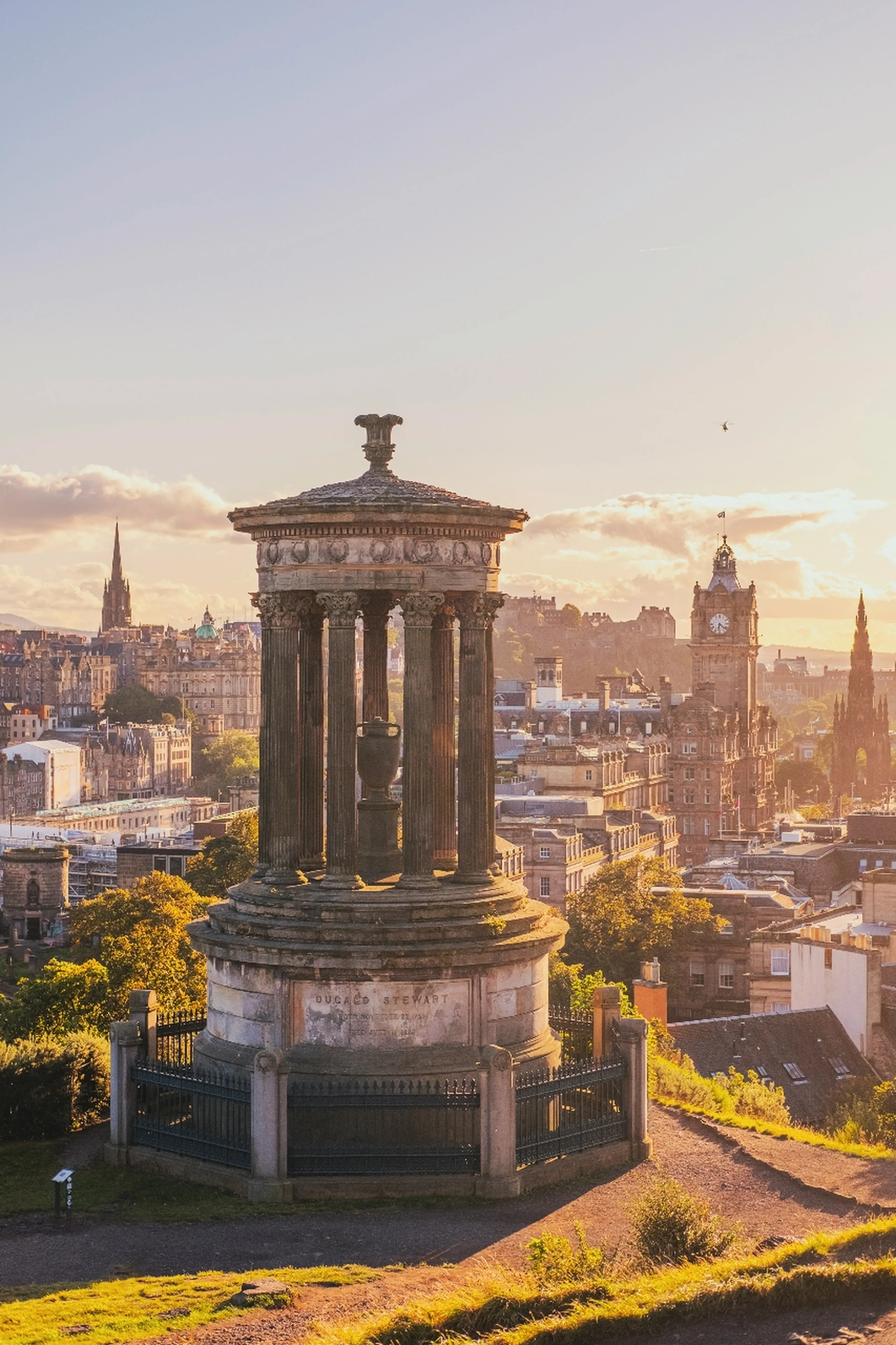 View of Dugald Stewart Monument on Calton Hill, Edinburgh, with cityscape and clock tower in the background during sunset.