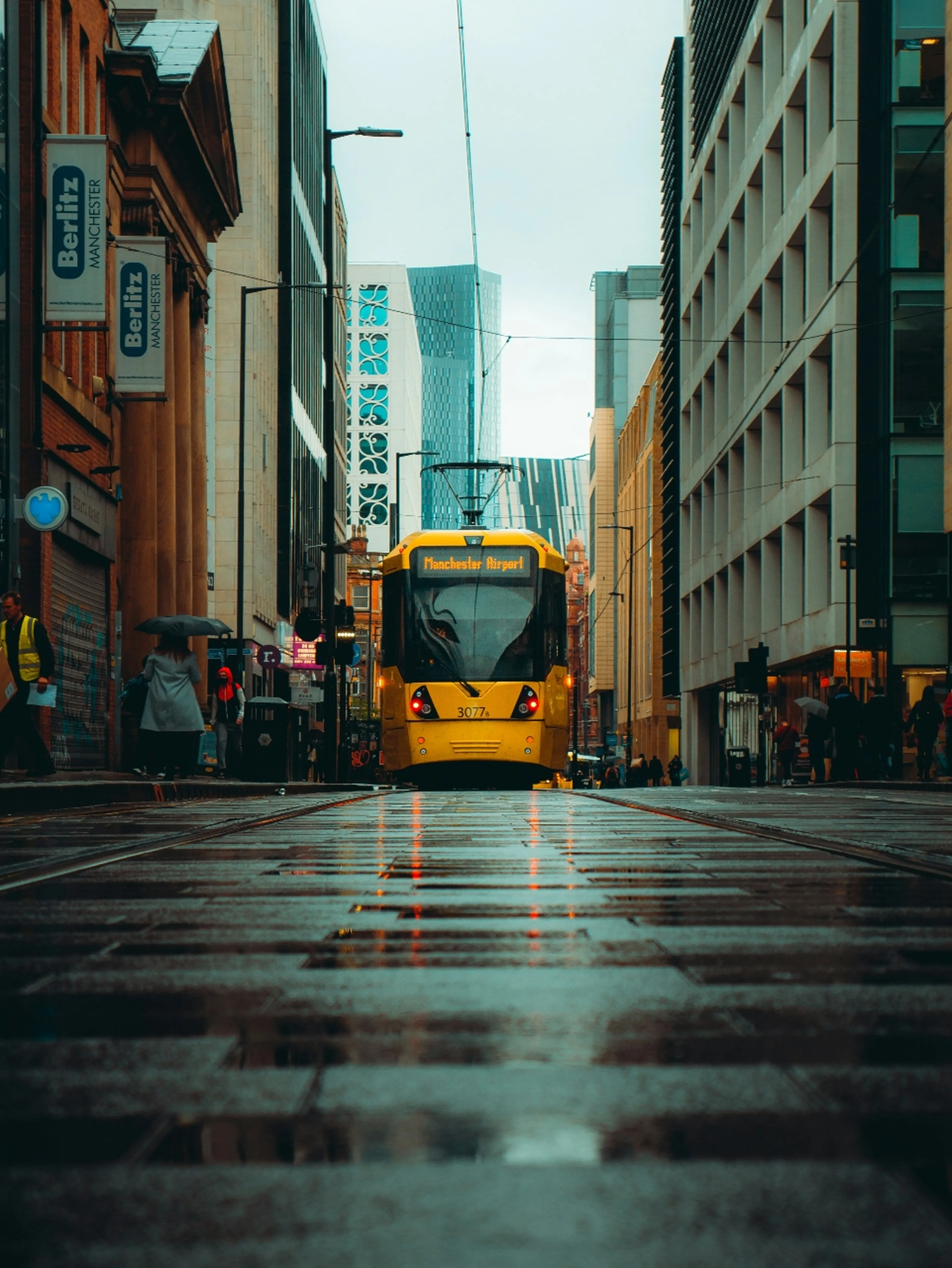 A yellow tram labeled "Manchester Airport" travels down a wet city street lined with tall buildings and pedestrians under umbrellas.