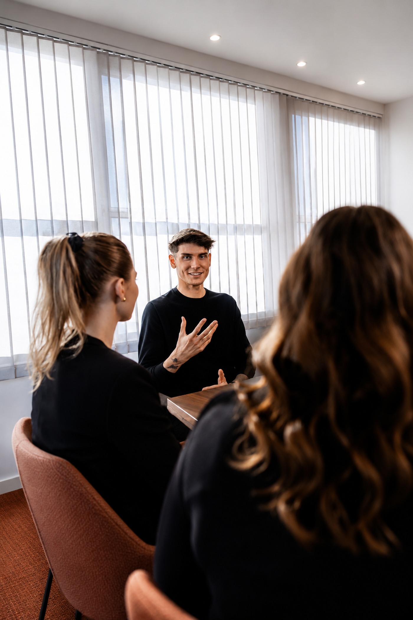 Ein Mann in einem schwarzen Hemd unterhält sich lebhaft mit zwei Frauen mit langen Haaren an einem Tisch in einem hell beleuchteten Raum mit Vertikaljalousien.
