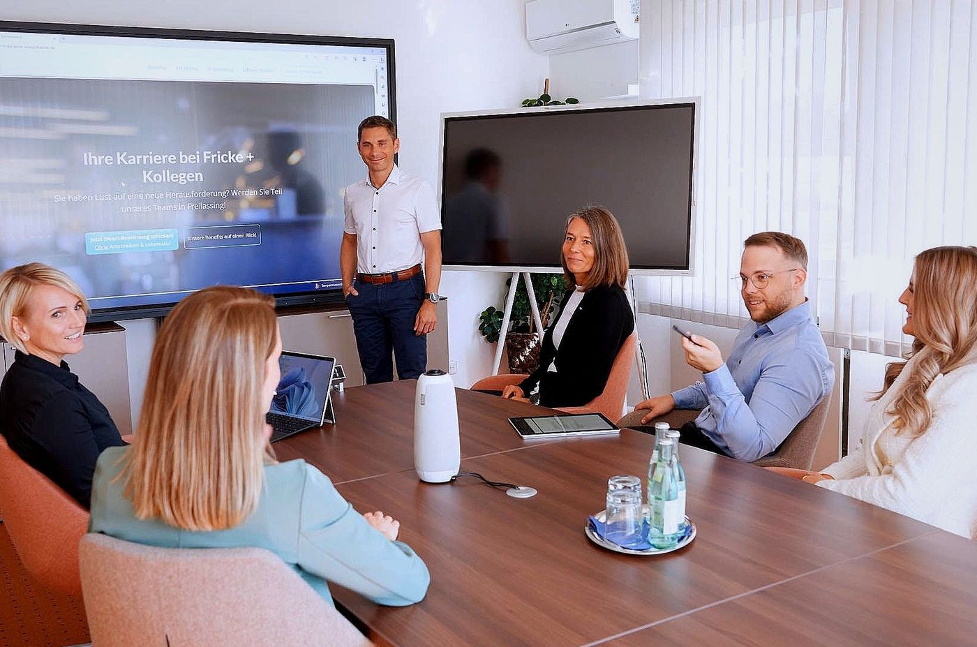 Teambesprechung in einem hellen Konferenzraum mit einer Präsentationsleinwand und sechs Personen, die um einen Tisch herum sitzen und Feedback geben.