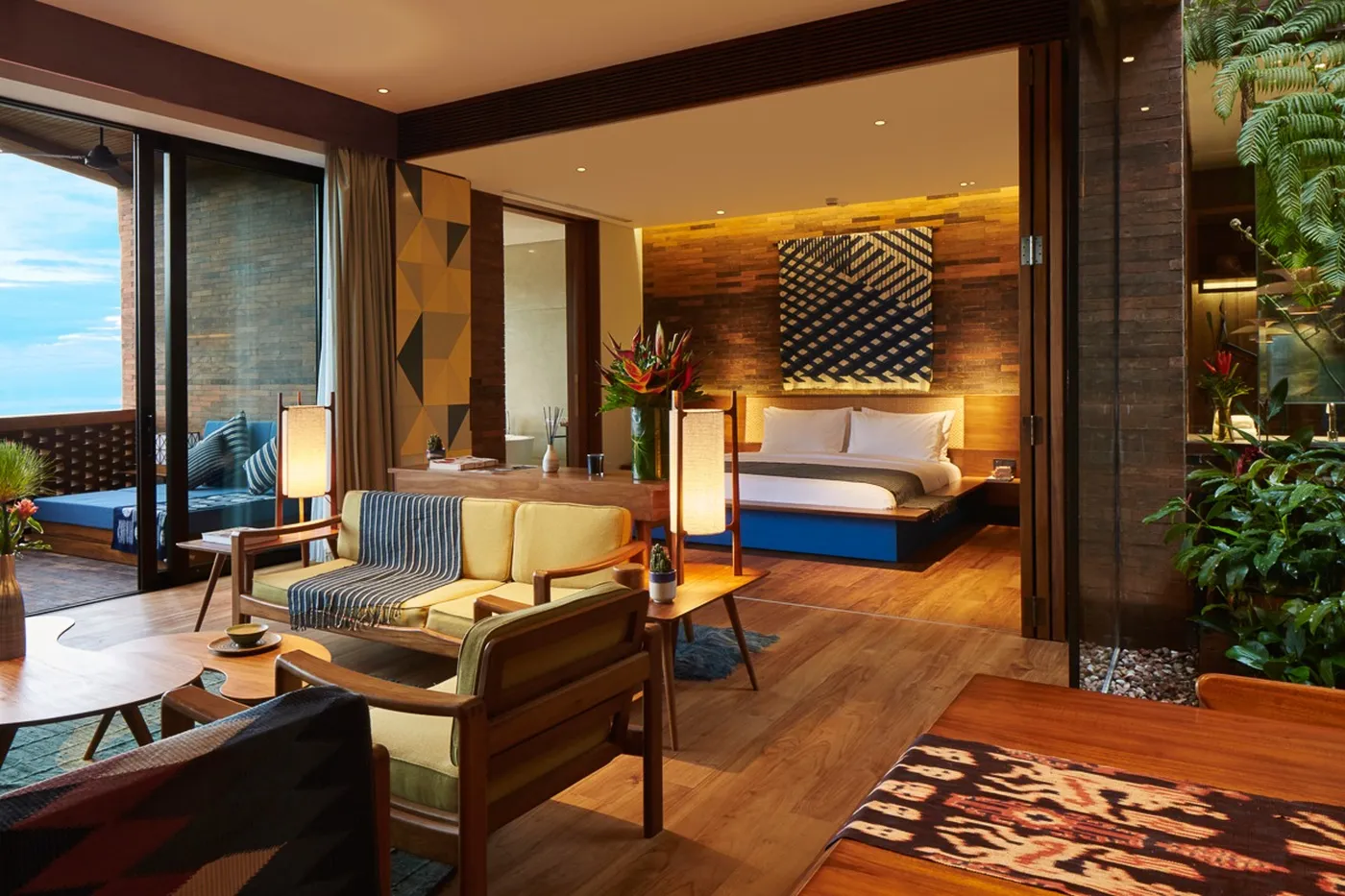 Luxurious hotel room with a king-sized bed, cozy seating area, wooden decor, large windows, and vibrant plants, creating a warm, inviting atmosphere.
