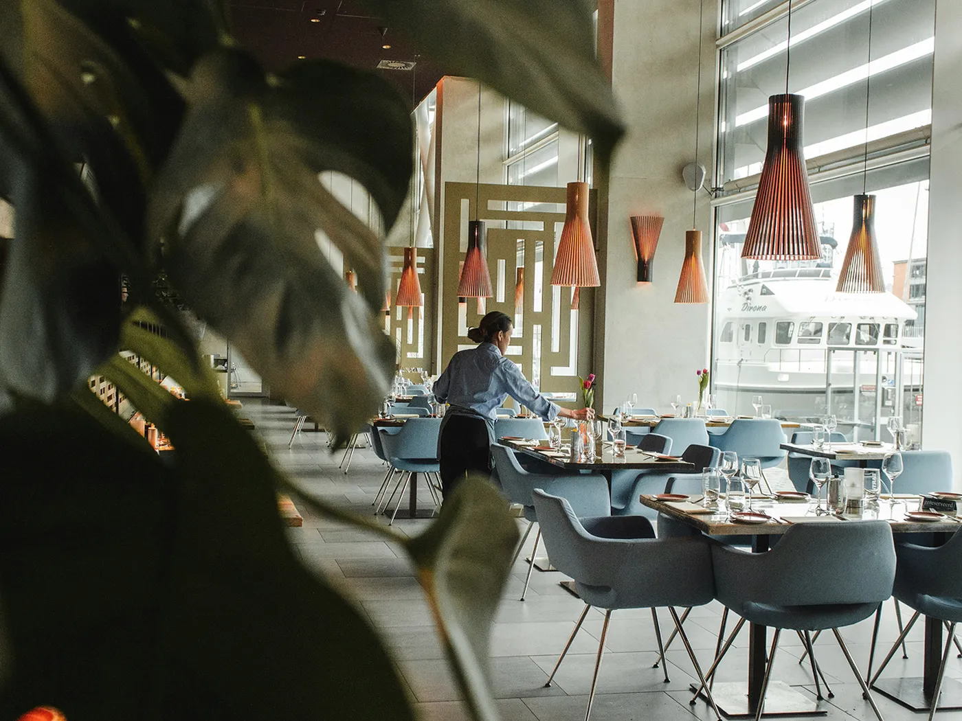 A person sets tables in a modern restaurant with blue chairs, large windows, and pendant lights. A plant is visible in the foreground.