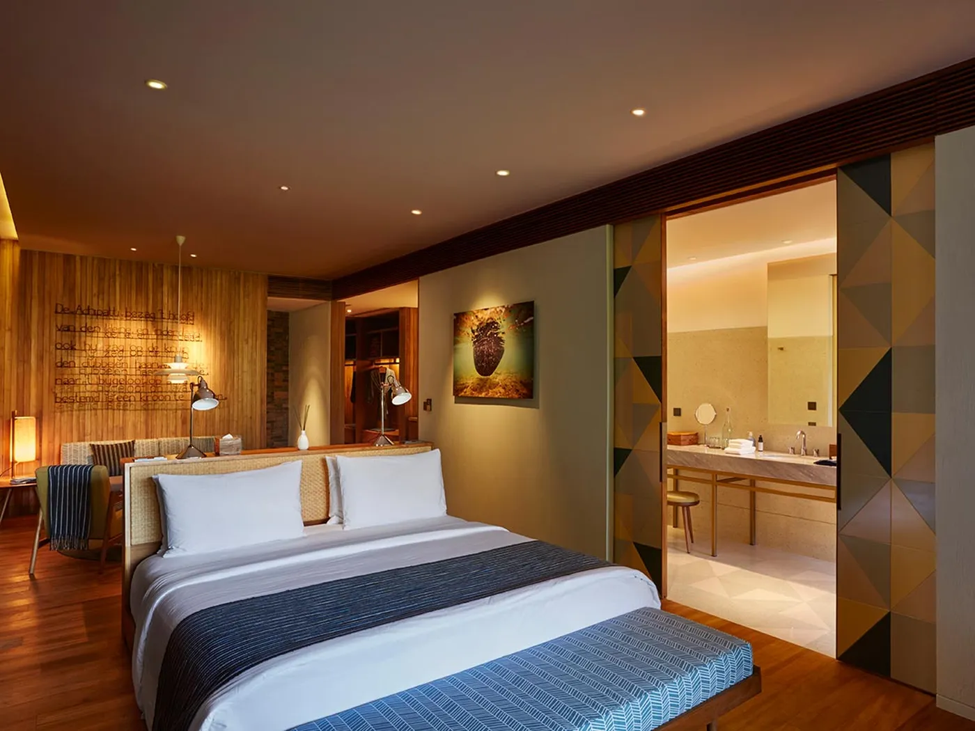 Modern hotel room with a double bed, decorative wall panel, and open view into a luxurious bathroom with a large mirror and vanity.