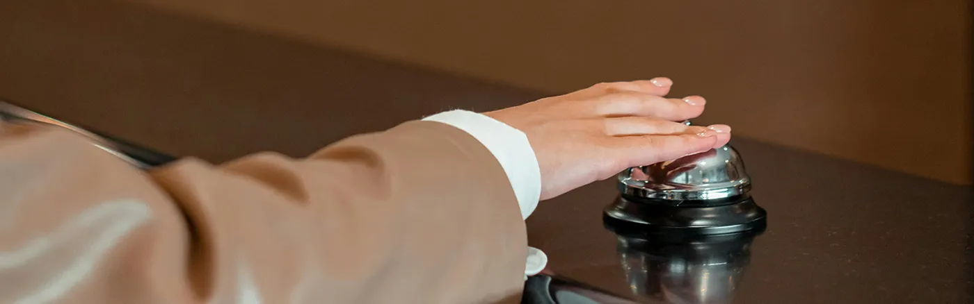 A person in a brown suit ringing a silver service bell on a dark countertop.