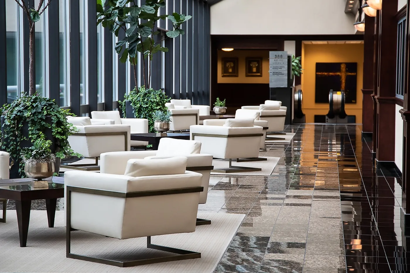 Modern lobby with white armchairs, glass tables, and potted plants along a polished floor, lit by natural light from large windows.