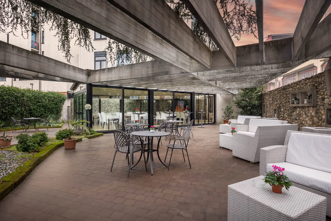 Outdoor patio with white sofas, round tables, and potted plants under a modern pergola. Glass-walled building in the background.