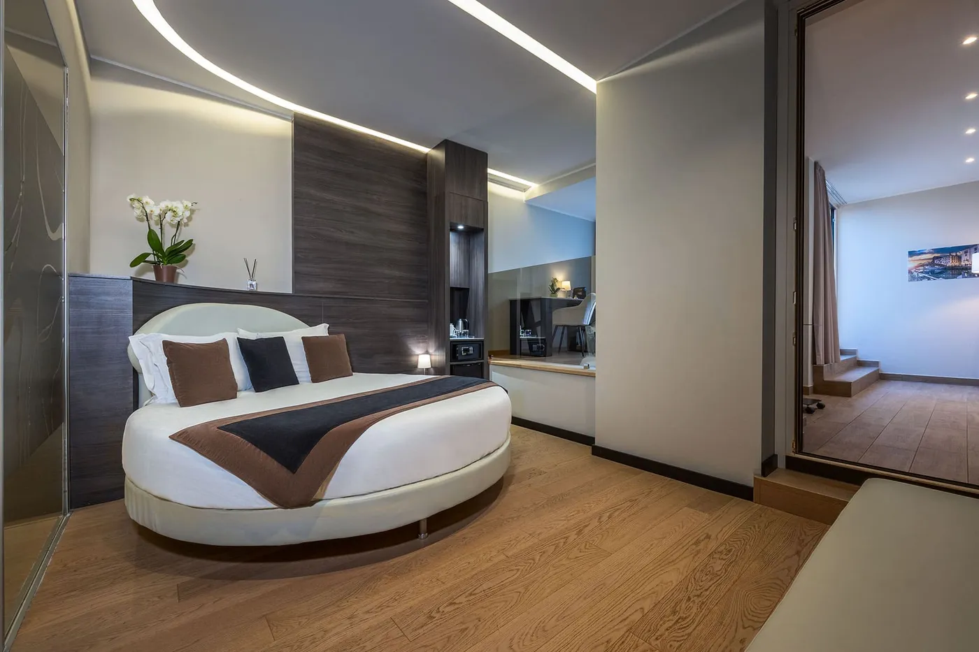 Modern hotel room with a round bed, brown and white bedding, wooden floors, large mirror, and minimalistic decor with soft lighting.