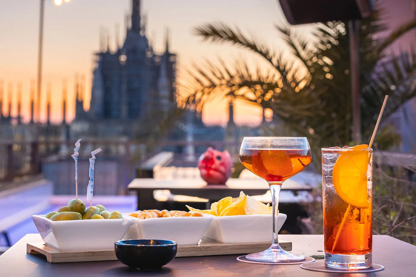 Cocktails and snacks on a rooftop table at sunset, with a blurred cityscape and palm in the background.