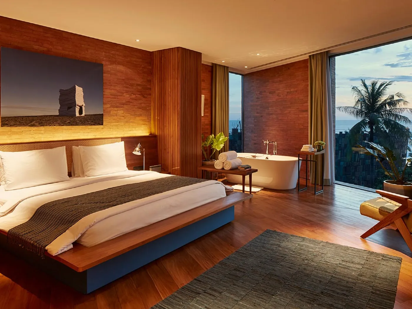 Luxurious hotel room with a large bed, freestanding bathtub, floor-to-ceiling windows, wooden accents, and a view of palm trees at sunset.
