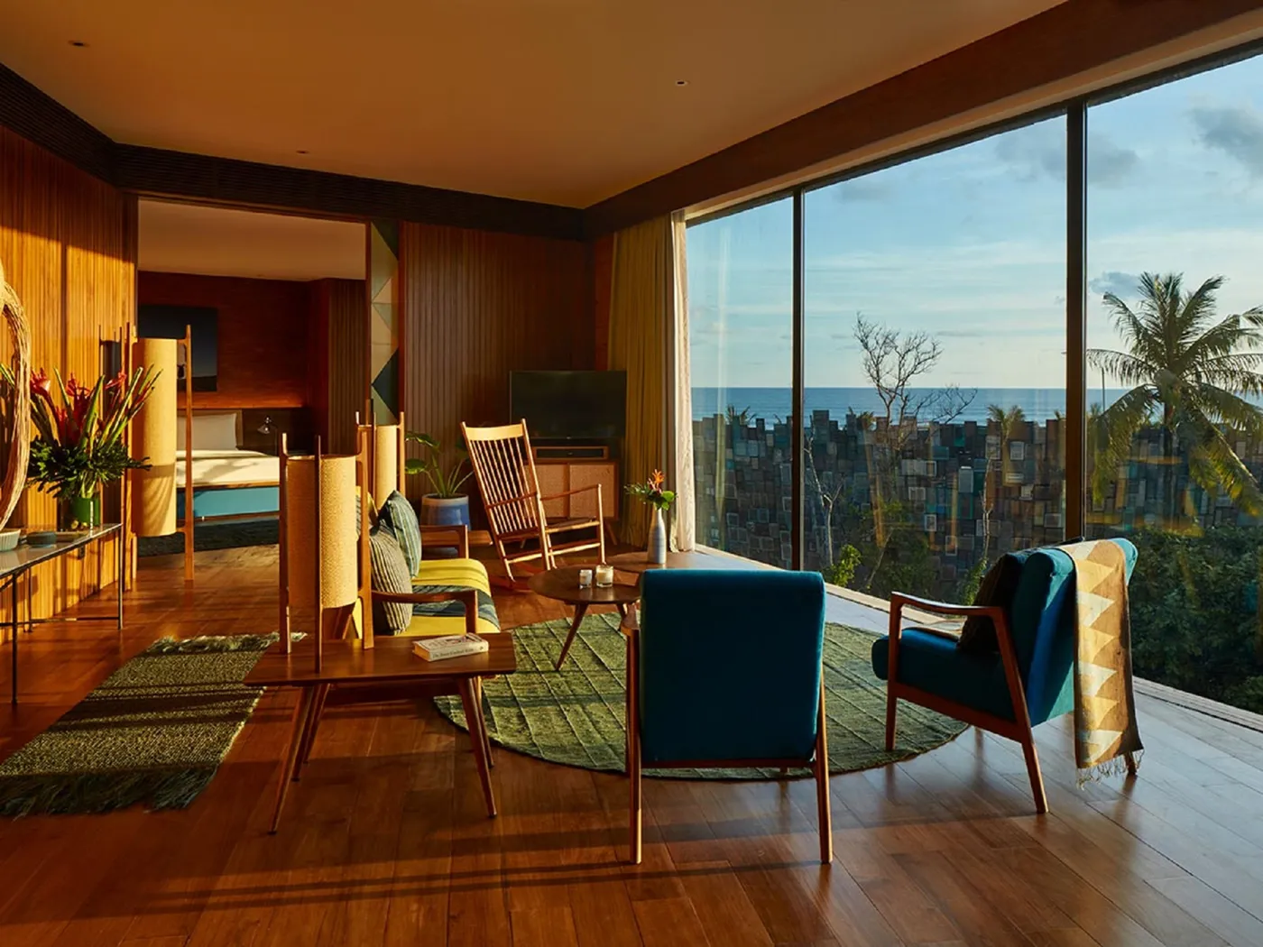 Spacious living room with wooden furniture, large windows offering an ocean view, and warm lighting. Tropical plants visible outside.