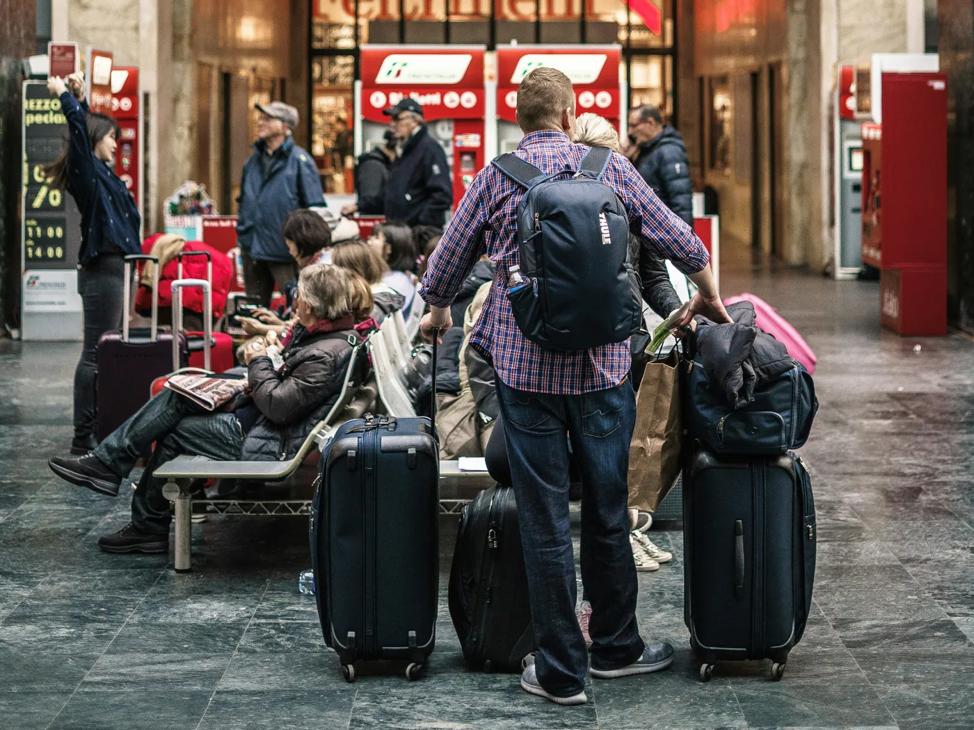 People with luggage at a busy train station, some seated and others standing, surrounded by ticket machines and signs.