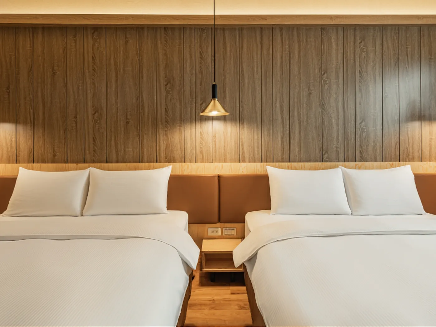 Two neatly made double beds with white linens in a modern hotel room, featuring a wooden accent wall and a hanging pendant light.