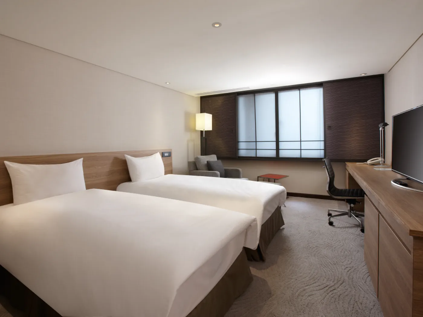 Modern hotel room with two twin beds, a desk with a chair, a flat-screen TV, an armchair, and a floor lamp by a large window with blinds.