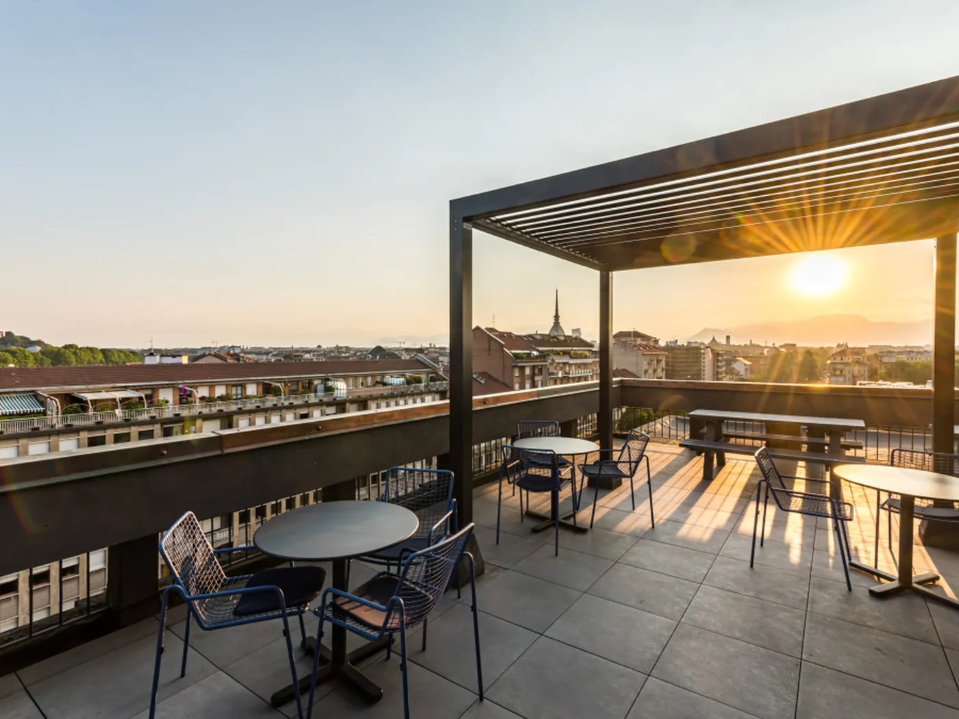 Rooftop terrace with tables and chairs, overlooking a cityscape at sunset. Sunlight casts long shadows on the tiled floor.