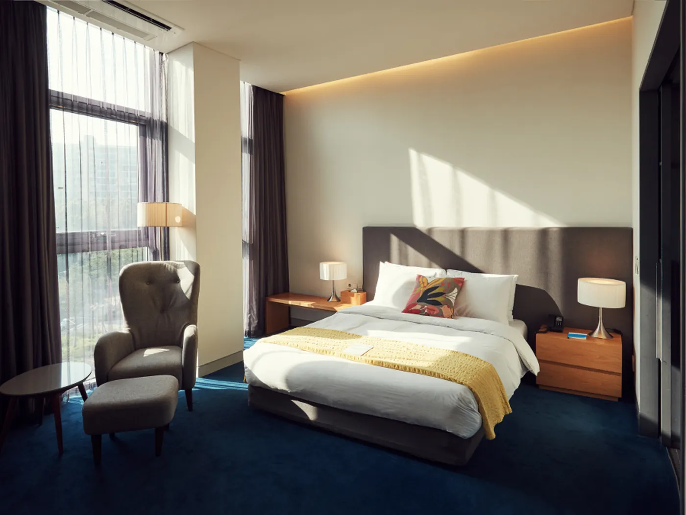 A modern hotel room with a large bed, yellow throw, armchair, side tables, lamps, and large windows with sheer curtains.