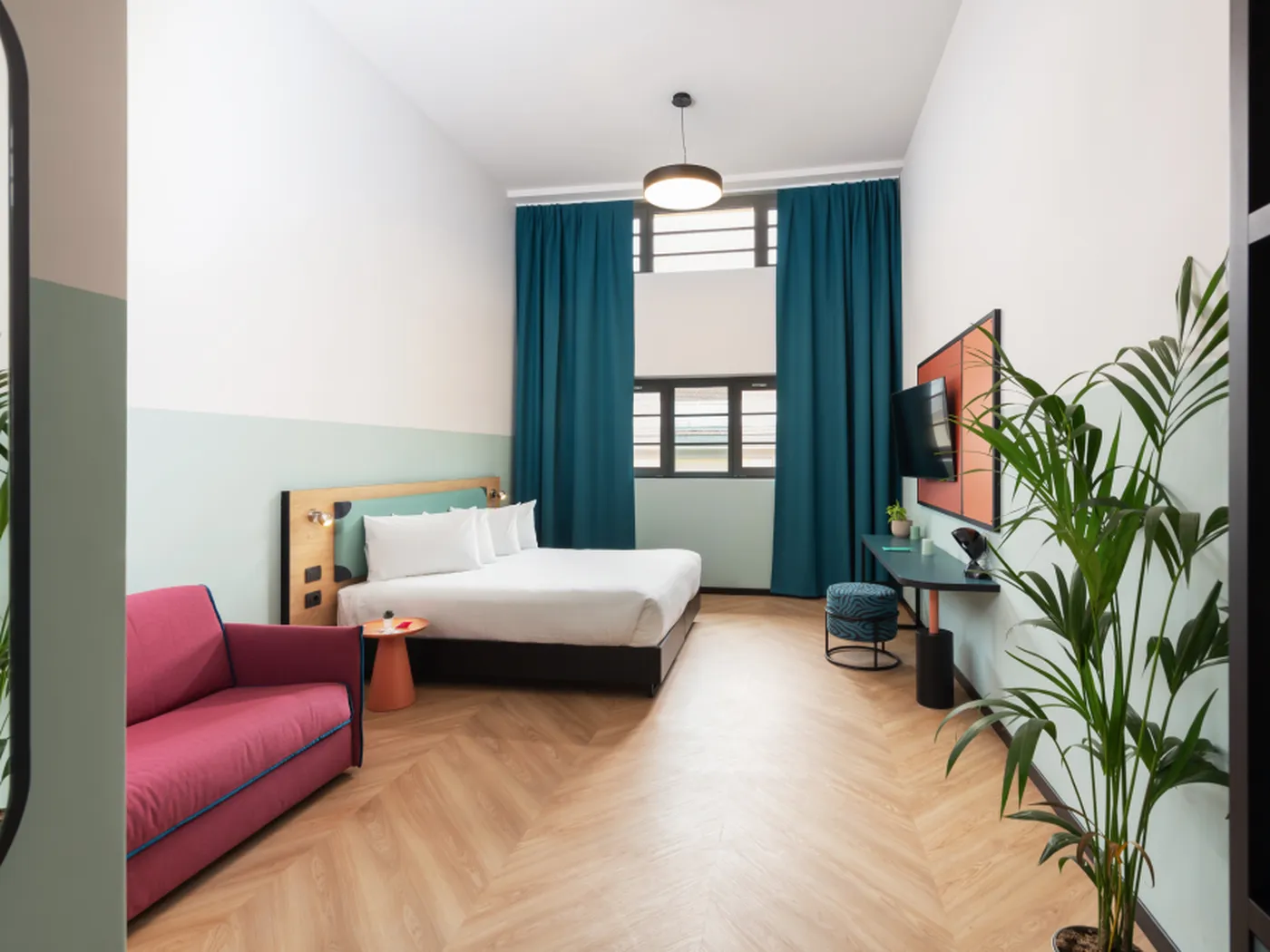 Modern hotel room with a large bed, pink sofa, teal curtains, wall-mounted TV, and a potted plant on wooden flooring.