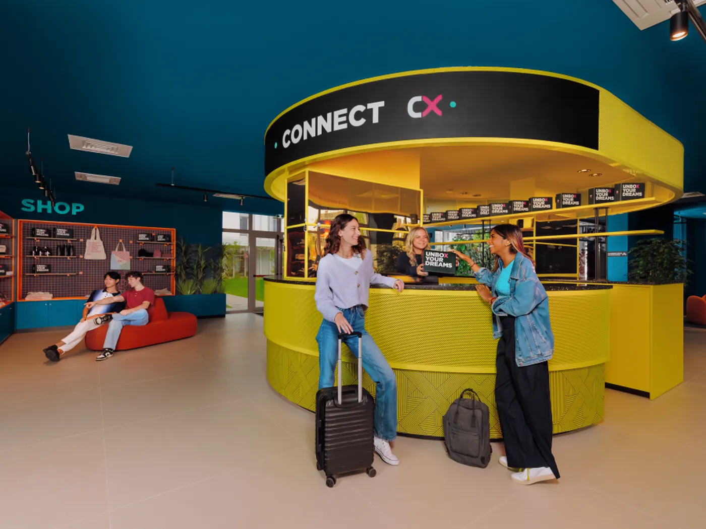 People socializing at a vibrant, modern cafe with luggage in a colorful interior. A couple sits on a red bench in the background.