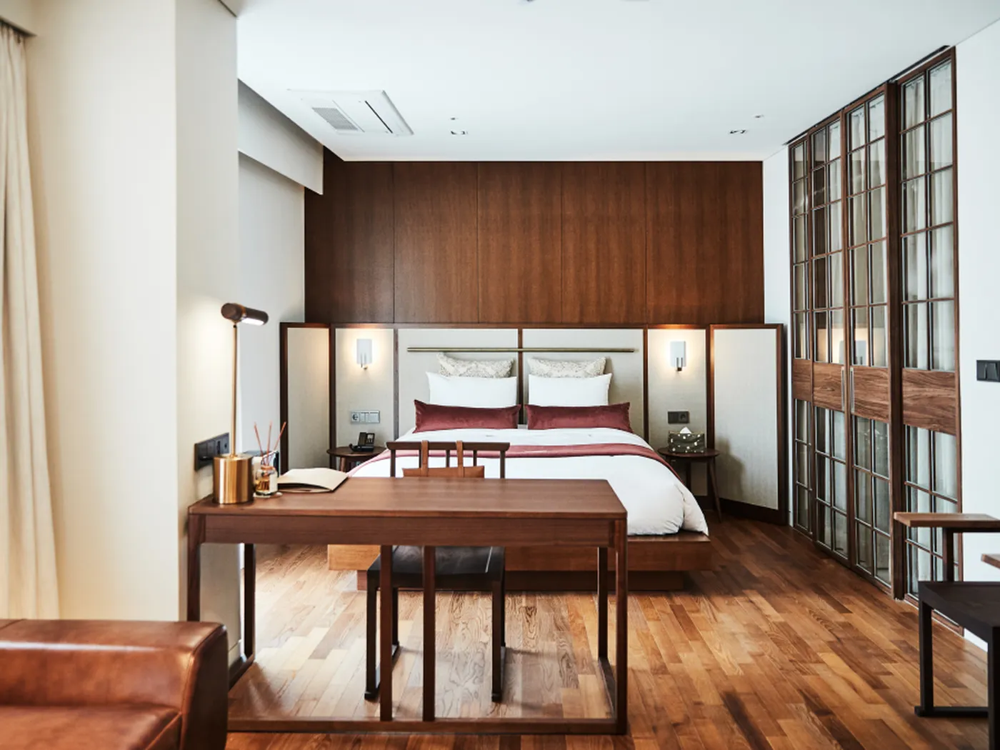 Modern hotel room with a wooden floor, double bed, desk, and floor-to-ceiling windows. Warm lighting and minimalist decor create a cozy ambiance.