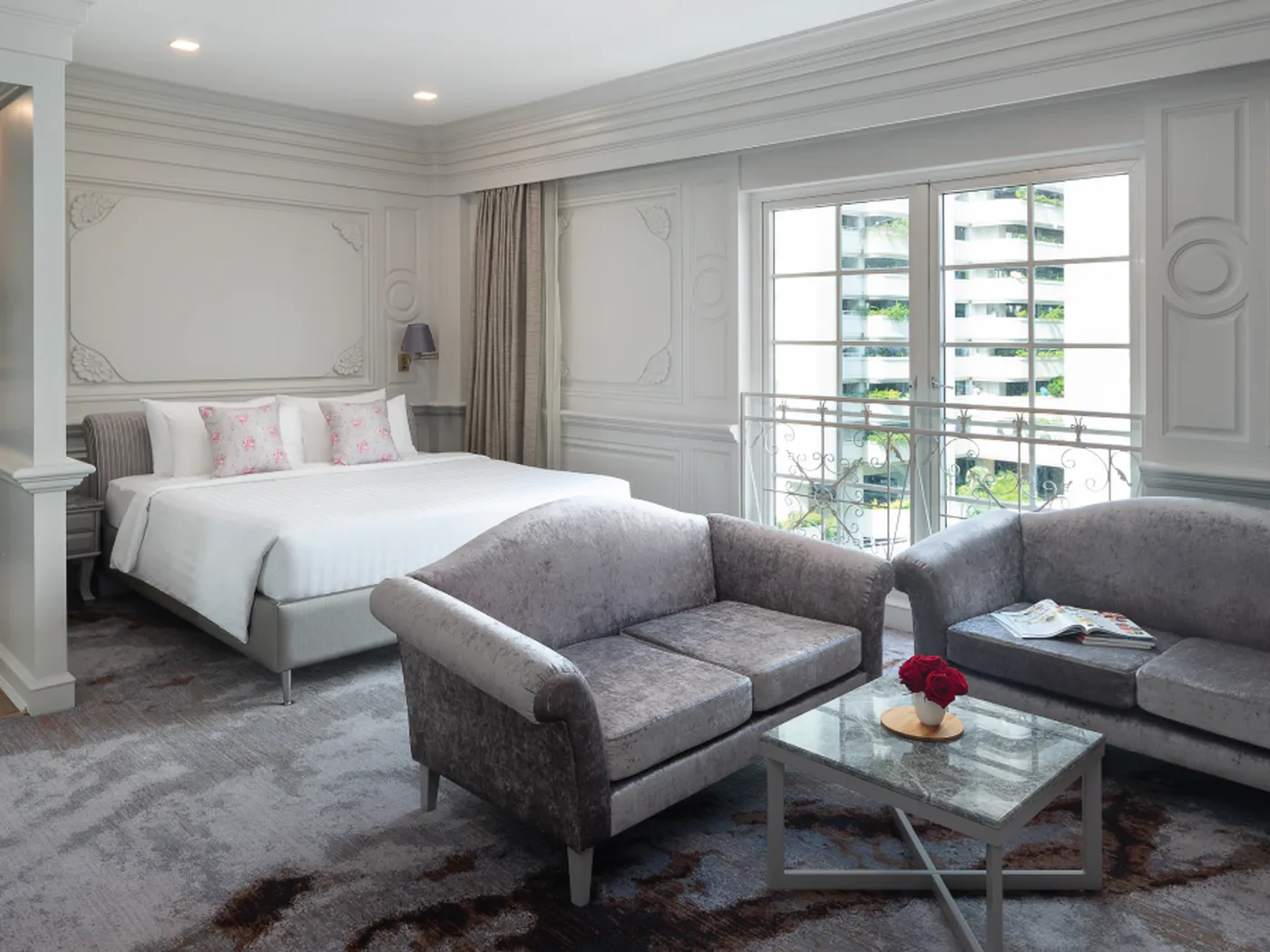 Elegant hotel room with a king-sized bed, two plush gray sofas, a glass coffee table with red flowers, and large windows with a city view.