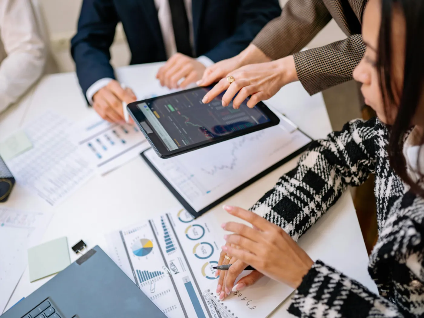 People in business attire review charts and graphs on paper and a tablet at a table, engaging in a collaborative discussion.