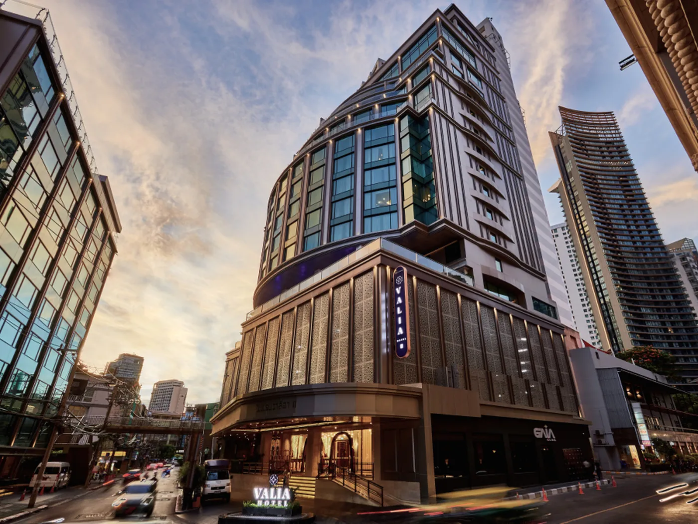 Modern high-rise building with glass facade under a blue sky with clouds. Street view includes nearby buildings and a road in the foreground.