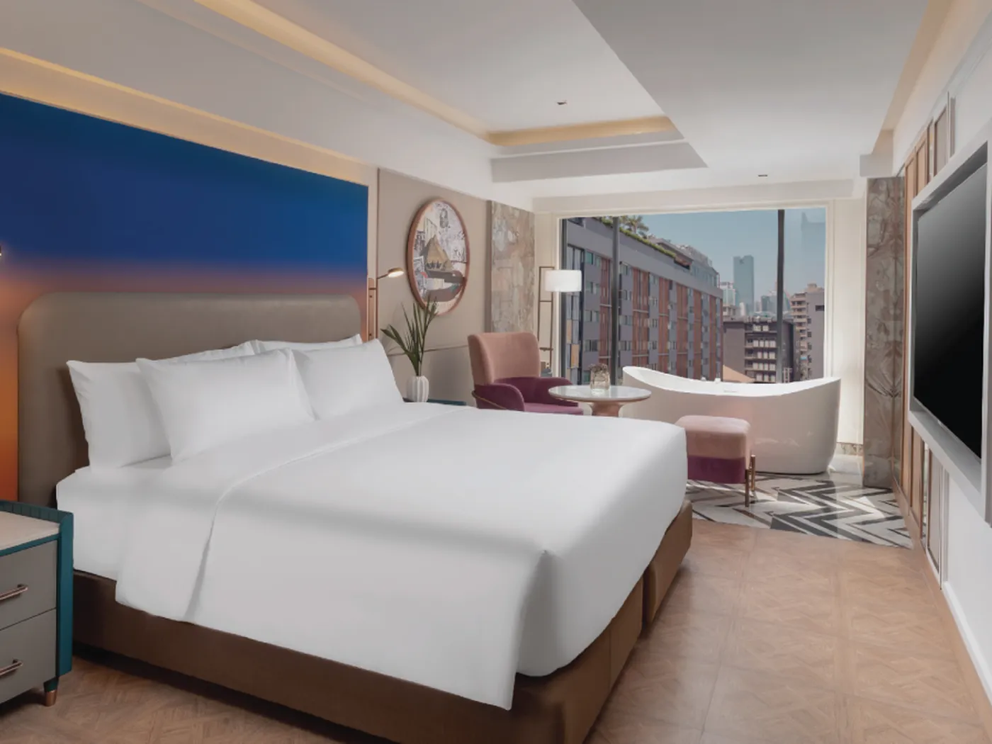 Modern hotel room with a large bed, armchairs, a round table, and a freestanding bathtub by the window overlooking a cityscape.