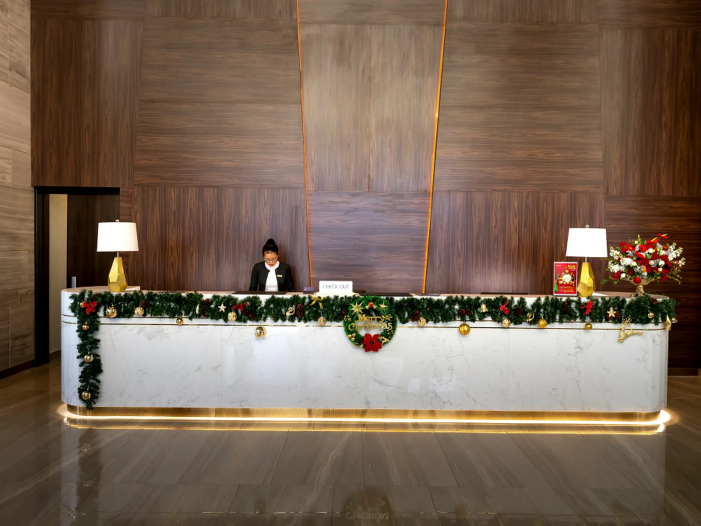 Hotel reception desk adorned with Christmas garlands and ornaments, flanked by lamps and a floral arrangement, with a staff member standing behind.
