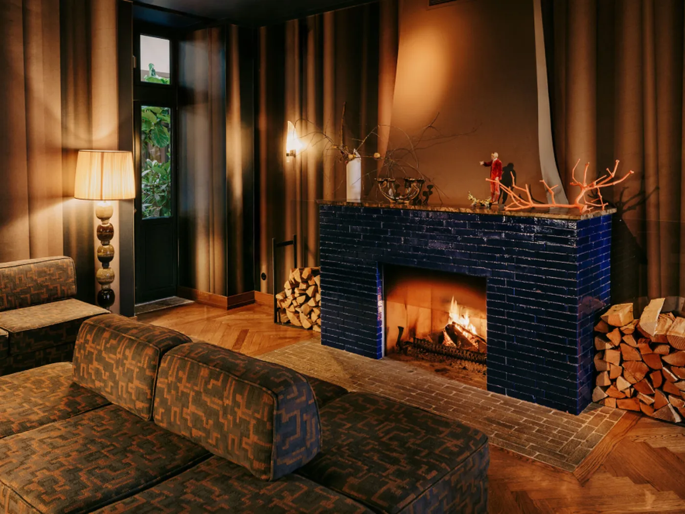 Cozy living room with a lit fireplace, blue-tiled mantel, patterned sofas, a stack of firewood, and a tall lamp beside a window.