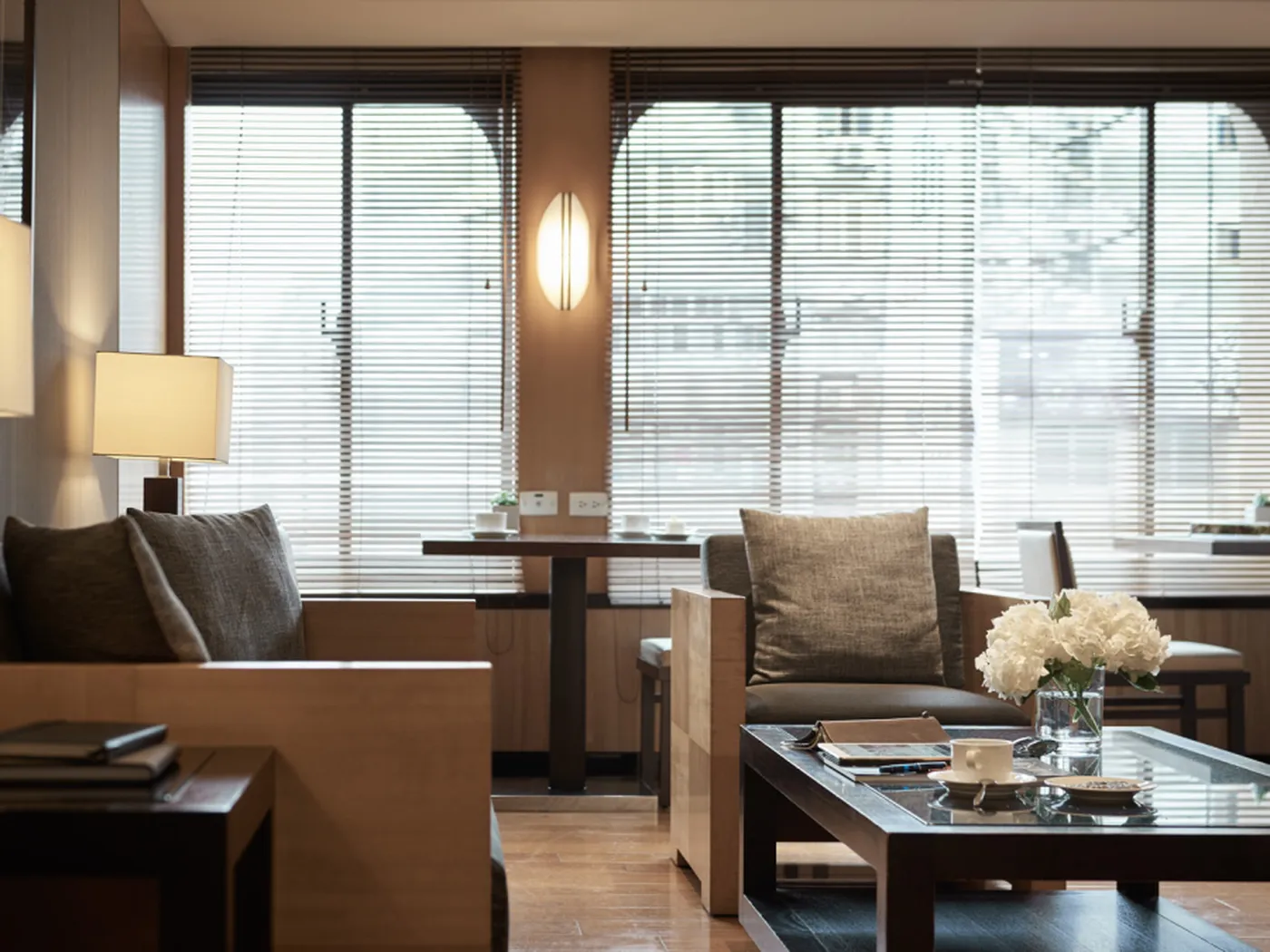 Modern living room with large windows, wooden furniture, gray cushions, a coffee table with books and flowers, and soft lighting.