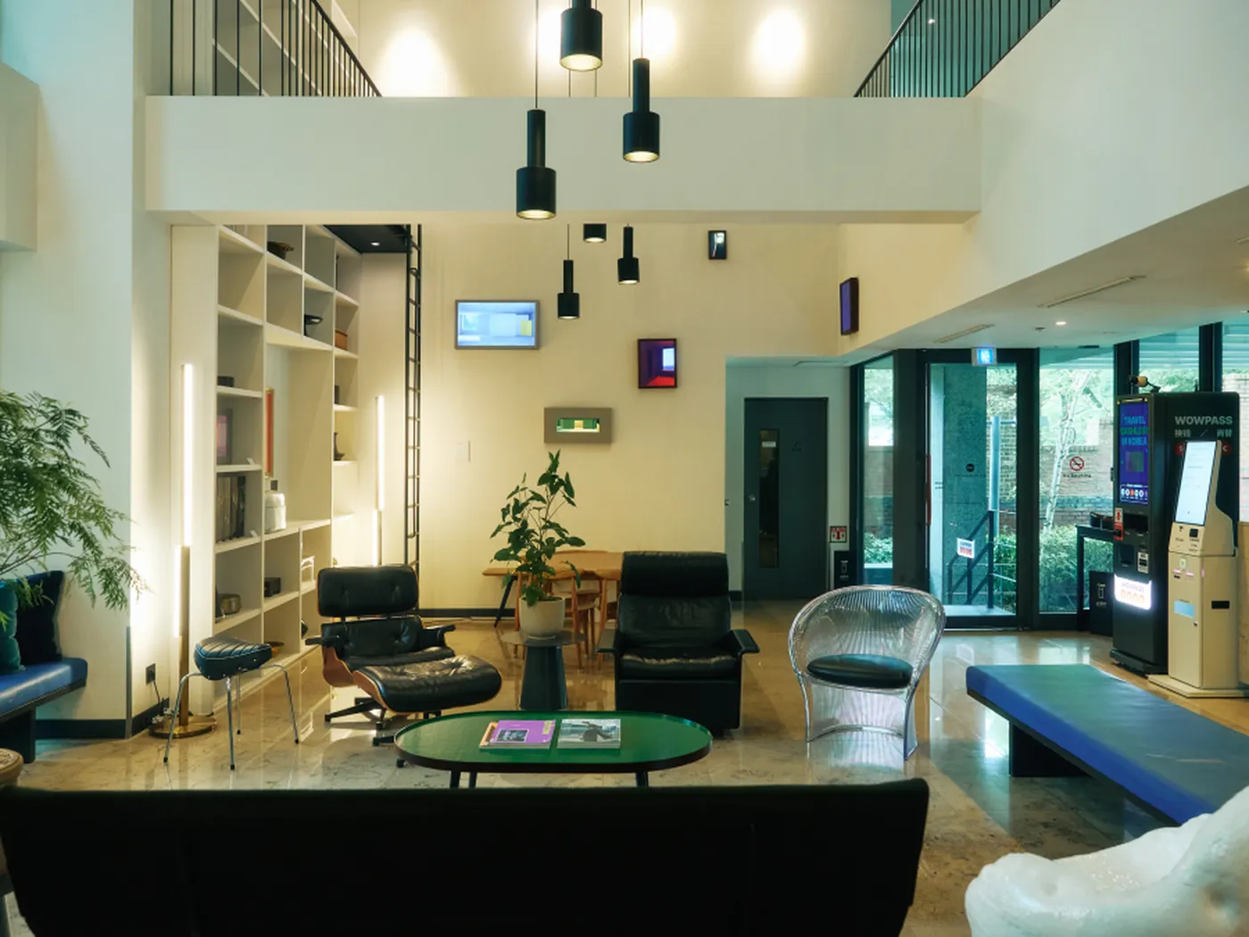 Modern lounge area with stylish seating, bookshelves, hanging lights, and a green coffee table. Large windows offer natural light and outdoor views.