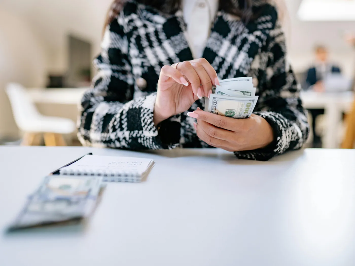 Person in a plaid jacket counting a stack of US dollar bills at a desk, with blurred office background.