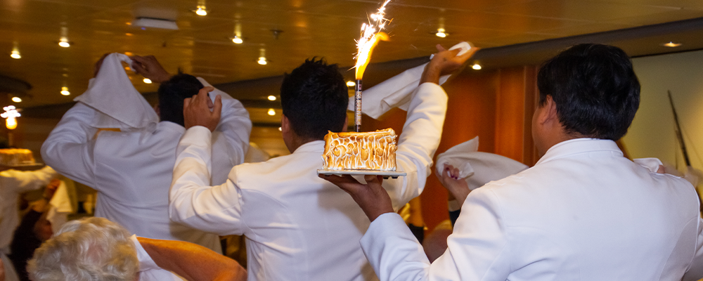 Waitstaff in white uniforms carry baked alaska with a sparkler through a restaurant