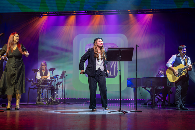Performers on stage with vibrant lighting; a singer at the center, musicians playing drums, guitar, and keyboard in the background.