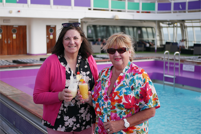 Anne & Yvonne smiling by a pool on the ship, holding tropical drinks.