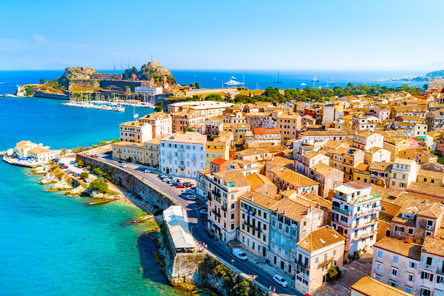 Aerial view of Corfu with colorful buildings, a fort on a hill, and a vibrant blue sea under a clear sky.