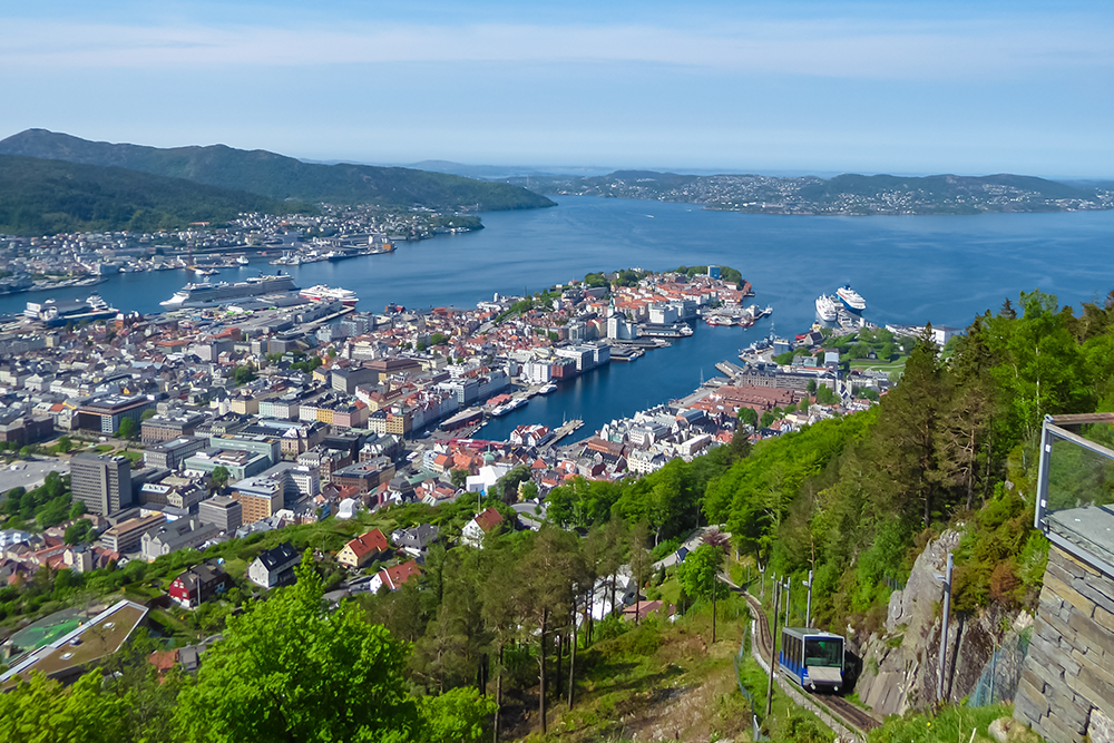 The Fløibanen funicular ascends Mount Fløyen, offering tourists on the viewing platform a spectacular panoramic vista of Bergen, Norway's cityscape, harbor, and fjords on a clear, sunny summer day.