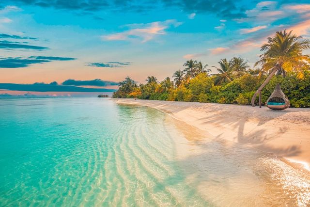 Tropical beach at sunset with clear turquoise water, white sand, lush palm trees, and a hanging wicker chair.