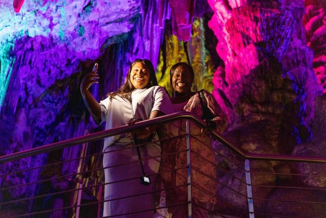Jacque & Tamara smile on a railing inside a cave, illuminated by colorful lights in pink, purple, and yellow hues.