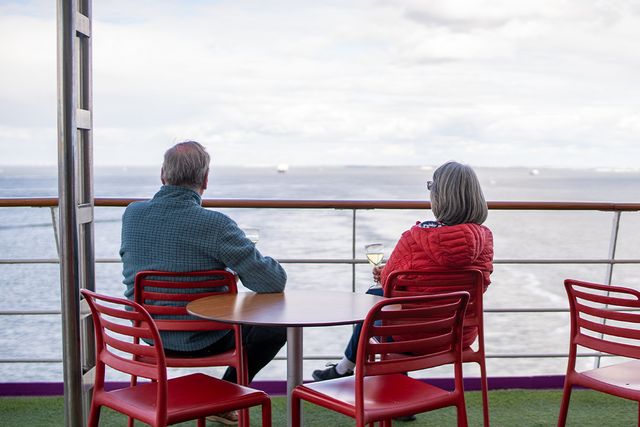 Two people sitting at a table on a ship's deck, looking out at the ocean, holding wine glasses.