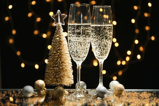 Two champagne glasses with bubbles, a small gold Christmas tree, and festive lights in the background on a reflective surface.