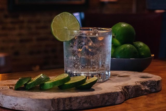 A glass of gin with a lime slice on the rim, surrounded by sliced limes on a wooden board, with whole limes in the background.