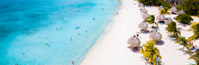 Aerial view of a beach in Curacao with turquoise water, white sand, people swimming, and straw umbrellas surrounded by palm trees.