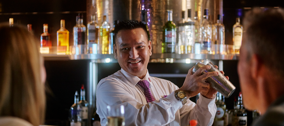 Bartender shaking a cocktail shaker, smiling at guest seated at a bar.