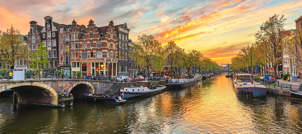 Sunset over an Amsterdam canal with boats and buildings lining the water, and a dramatic sky with vibrant clouds in Amsterdam, Netherlands.