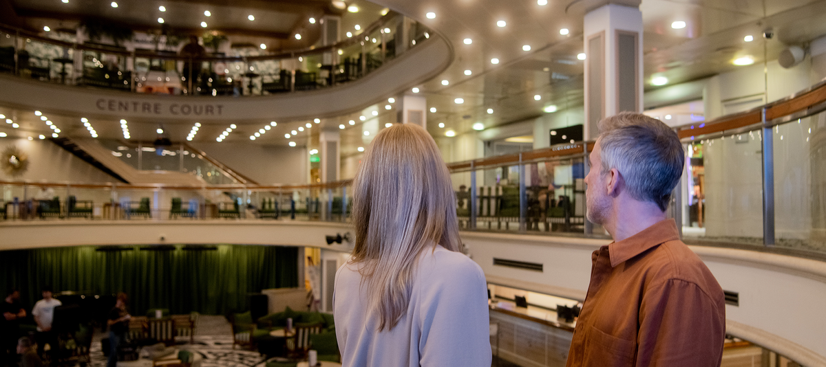 A man and woman overlook the spacious, Centre Court , surrounded by seating, and lit with numerous ceiling lights.