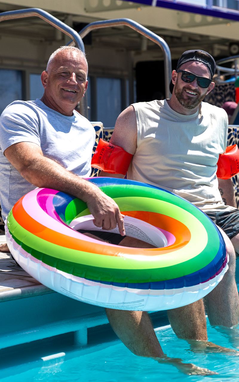 Graham & Lewis sitting by a pool; one holds a rainbow inflatable ring, both smiling. One wears water wings and sunglasses.