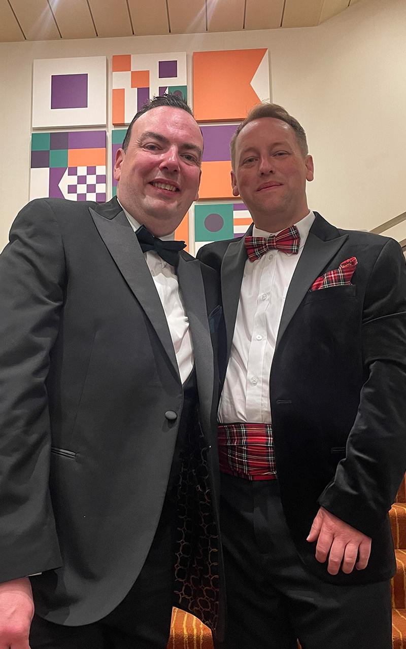 James & Damien in formal attire, one in a black tuxedo and the other in a velvet jacket with tartan accents, stand smiling in front of a geometric wall art.