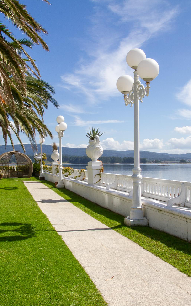 Pathway with white street lamps and palm trees beside a calm waterfront in Vigo, under a clear blue sky.